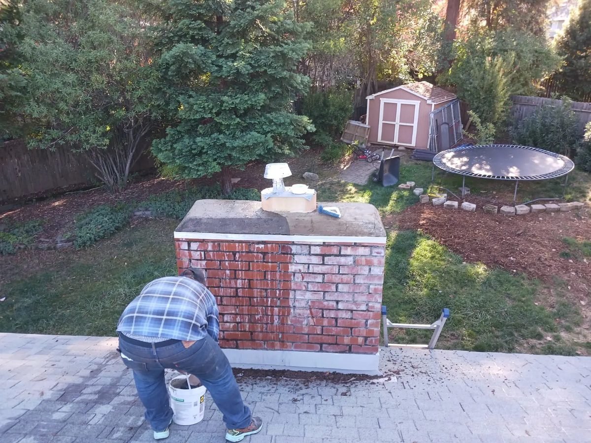 Mason working at roof level on a red brick chimney with a freshly cast concrete crown and repointed joints across the upper courses