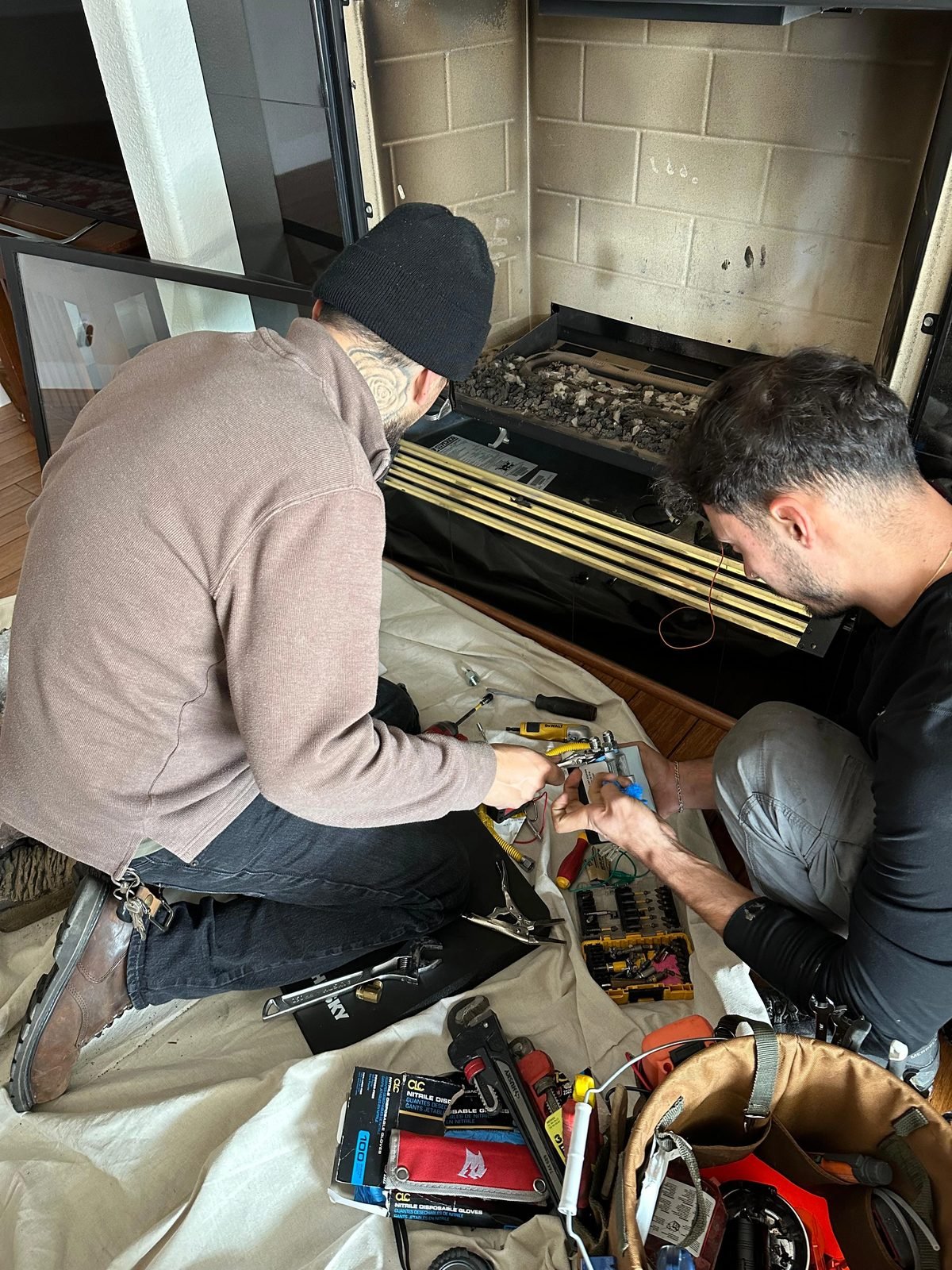 Two technicians kneeling on a drop cloth servicing the gas valve and pilot assembly of a fireplace, with the burner pan pulled forward and tools spread out
