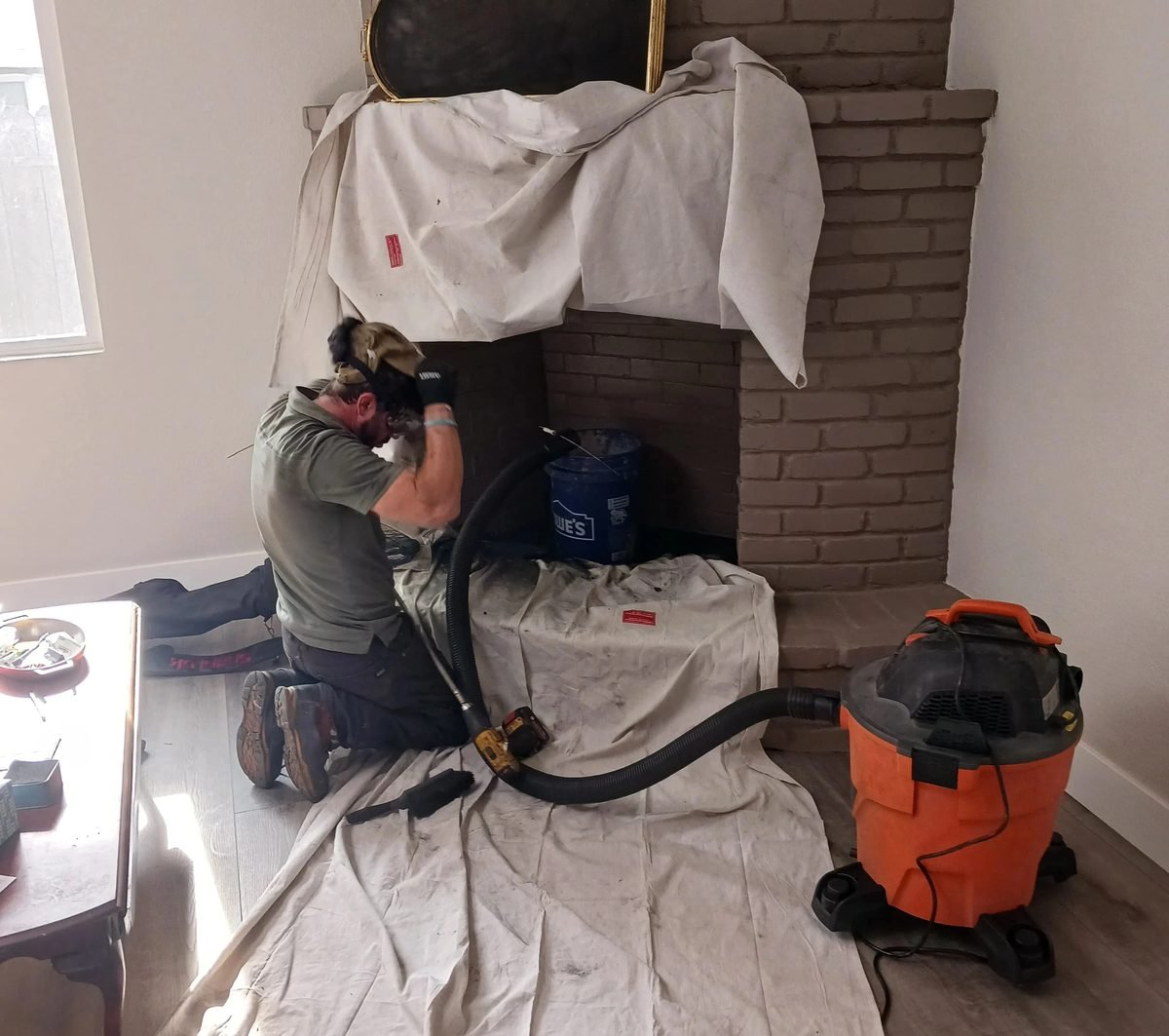 Technician kneeling on drop cloths at a painted brick fireplace, feeding a sweep hose past the damper with a shop vacuum running on the floor beside him