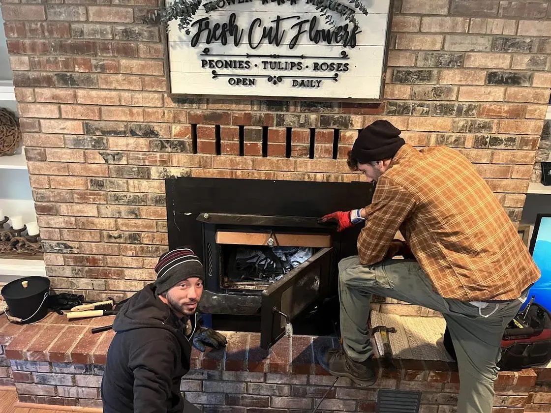 Two Brightwood technicians opening the doors of a fireplace insert set into a floor-to-ceiling brick chimney breast, with hand tools laid out on the hearth