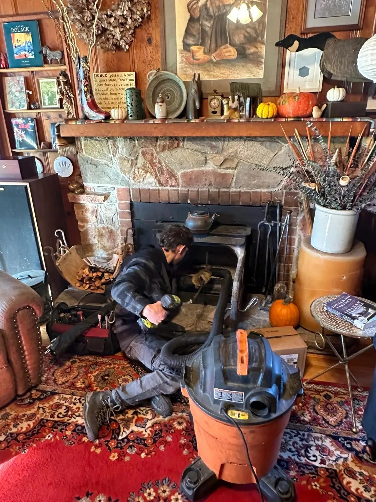 Brightwood technician kneeling at an open stone fireplace with a sealed HEPA vacuum running beside the hearth during a chimney sweep
